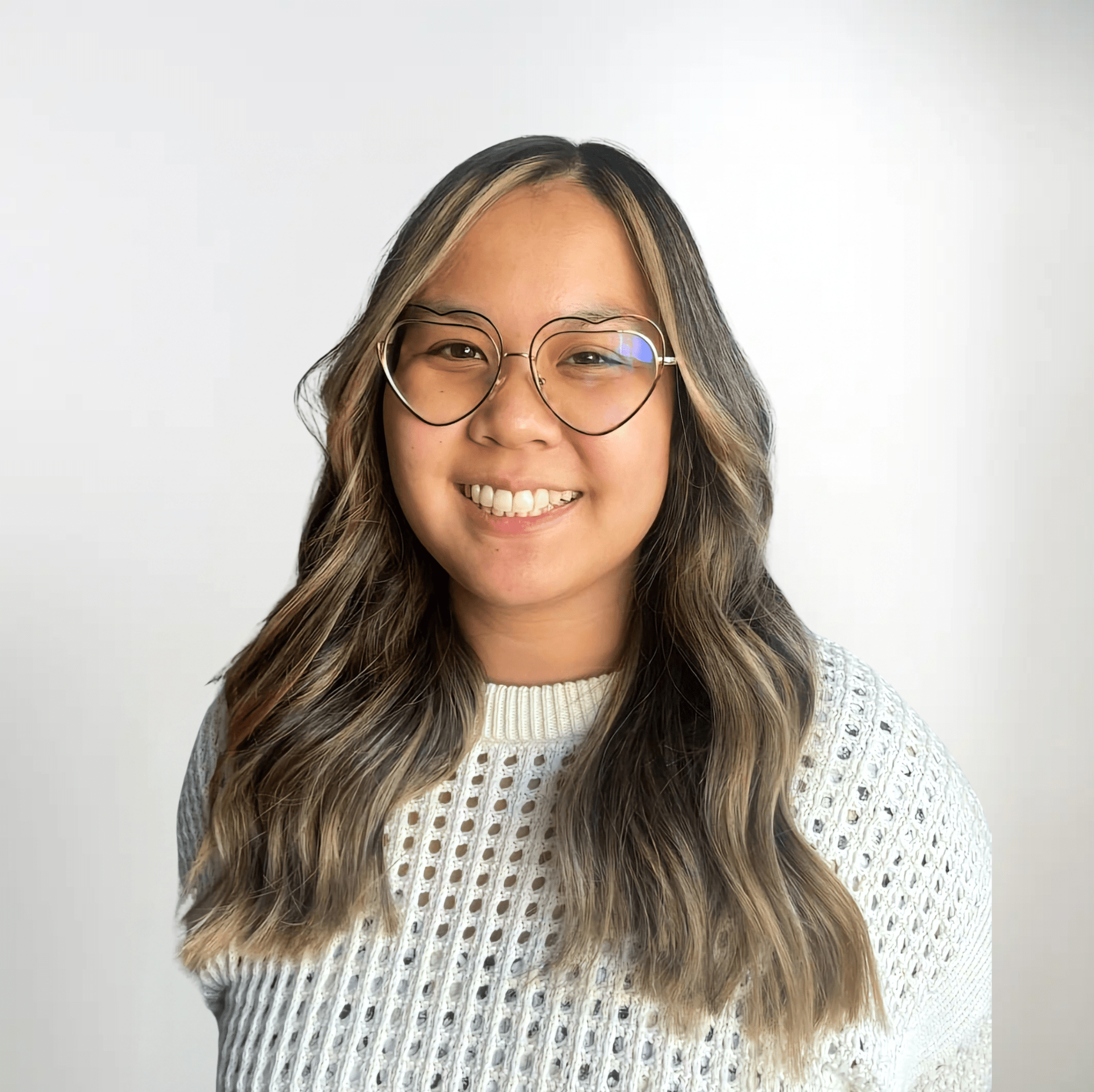 A headshot photo of a smiling woman with brown hair with blonde highlights wearing a white knit sweater and heart-shaped glasses.