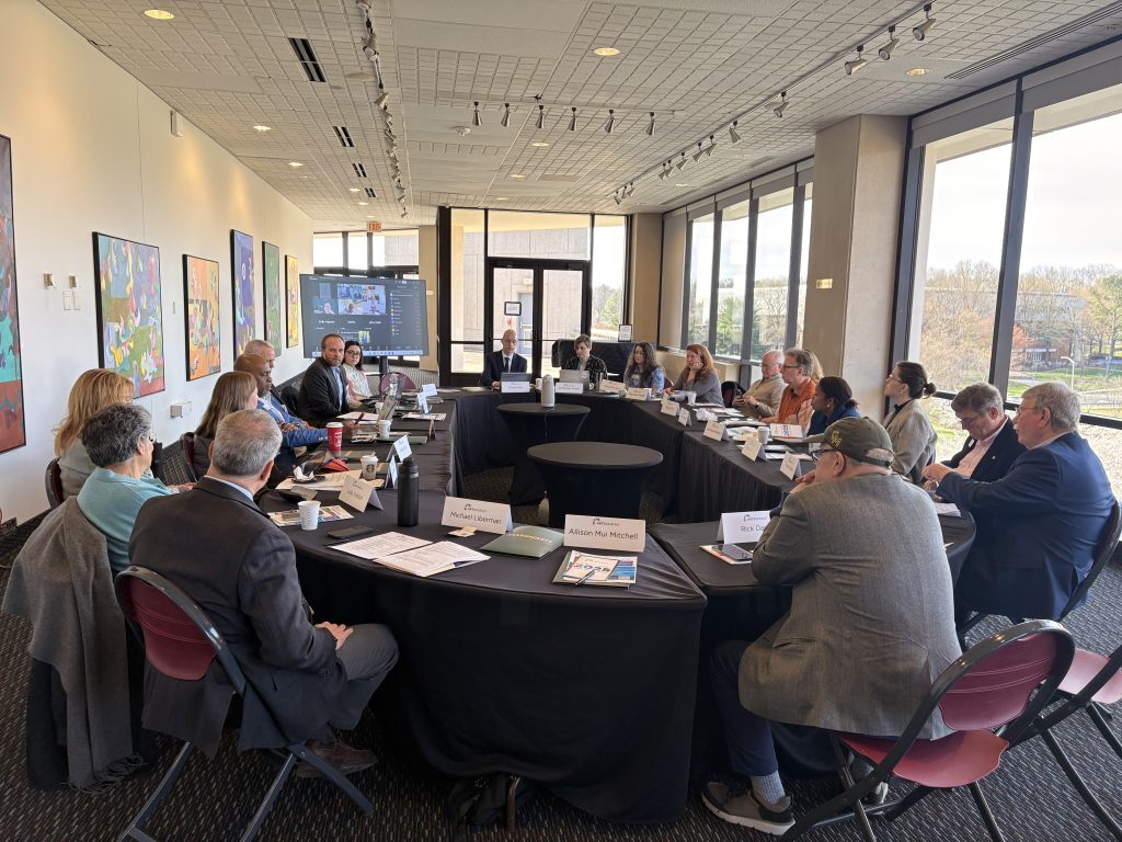 A photo of a large room with hung art along the left wall and floor-to-ceiling windows along the remaining walls surround a U-shaped table with people sitting all around it.
