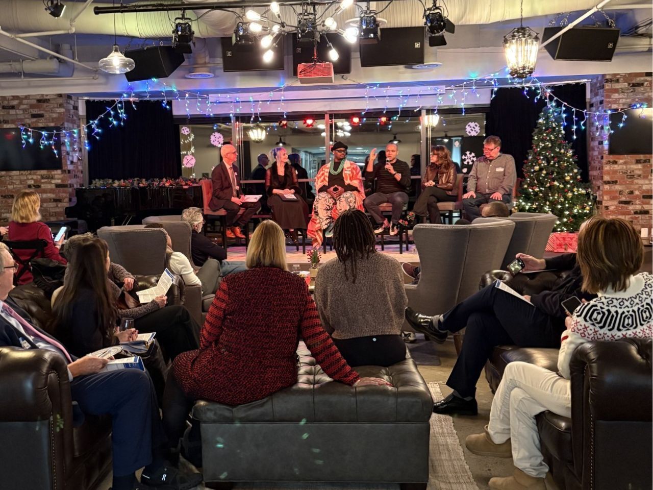 An audience seated in couches and armchairs face a seated panel on a low stage.