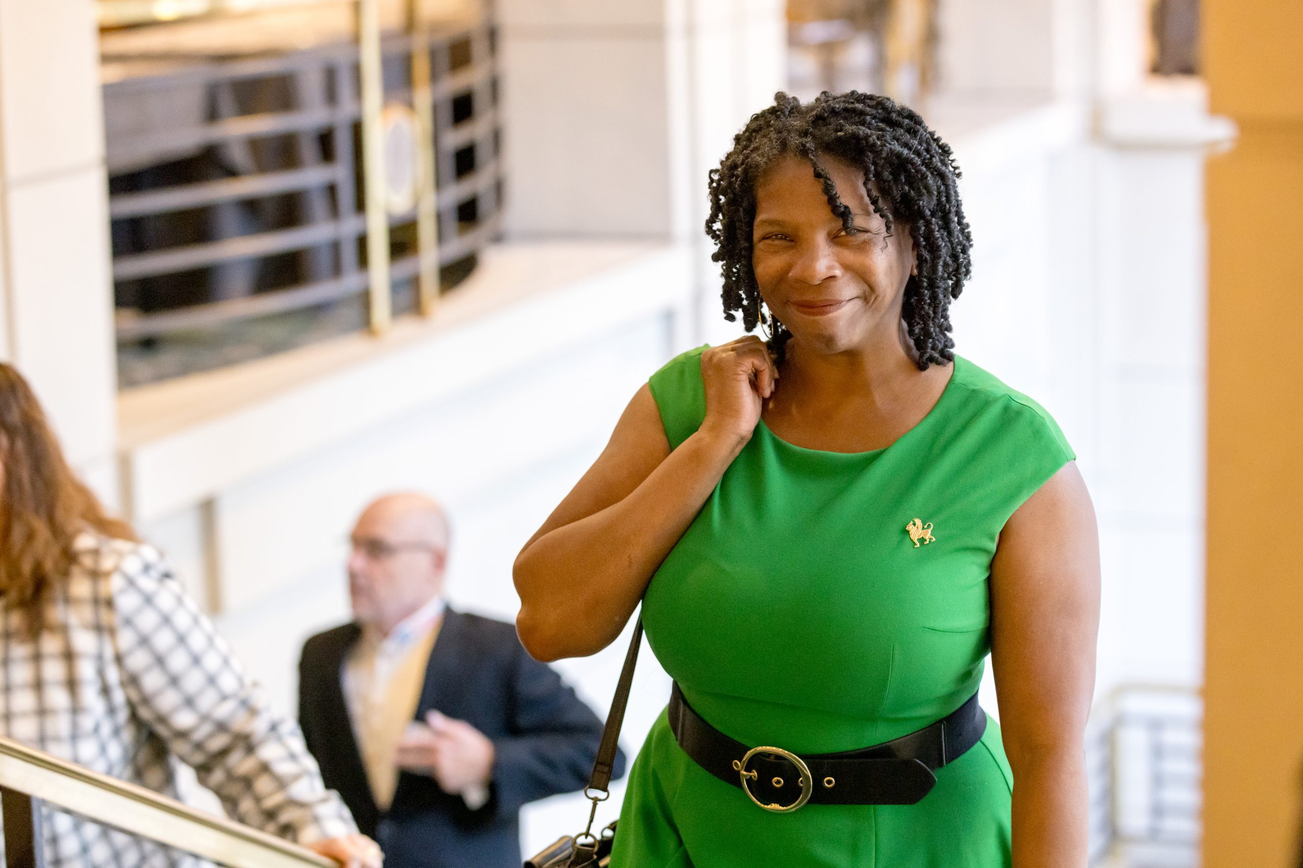 A black woman with chin-length twisted black hair in a green dress and black built looks up at the camera coming up a set of stairs.