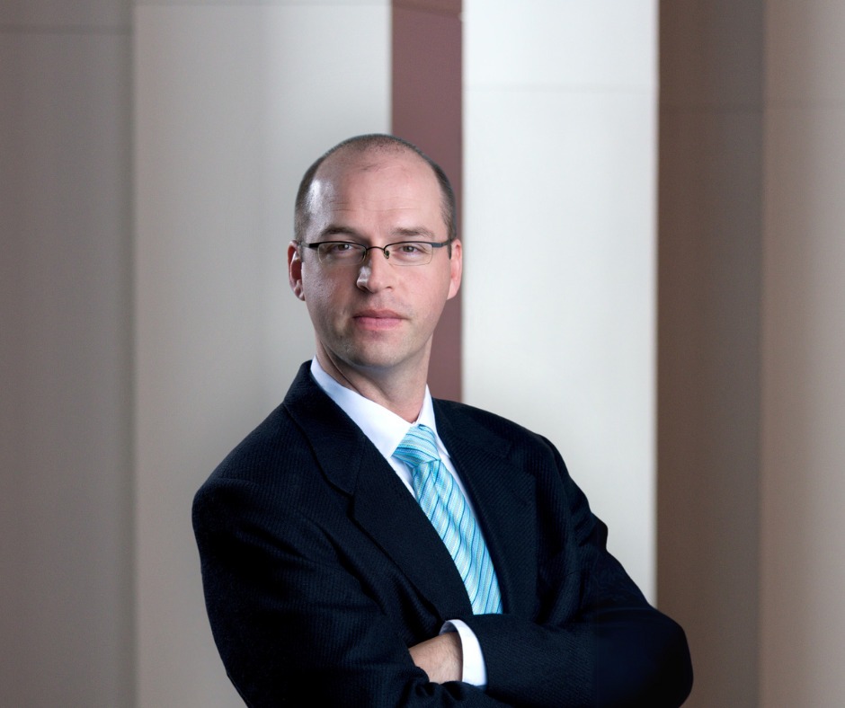 A man with close shaved dark hair and black wireframe glasses looks at the camera. The man wears a black suit