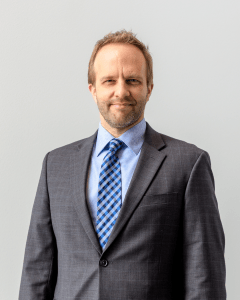 A man smiles in his headshot photo. He is wearing a light blue shirt and blue checkered tie with a gray suit jacket.