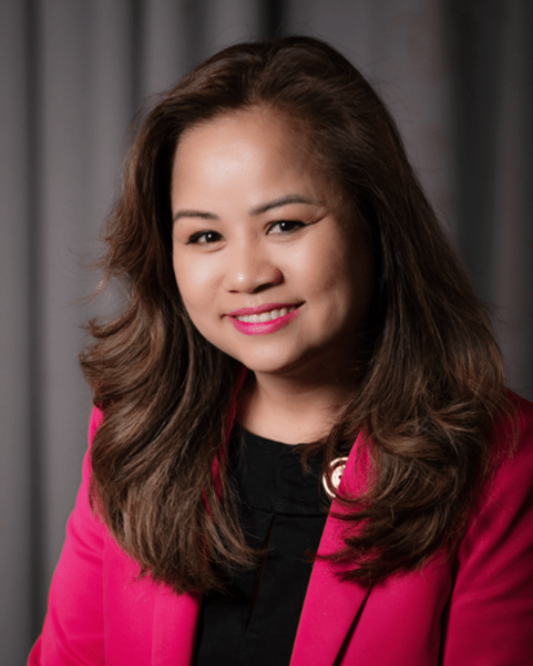 A woman smiles in a headshot photo. She has medium length dark brown hair and is wearing a black shirt with a dark pink blazer jacket.