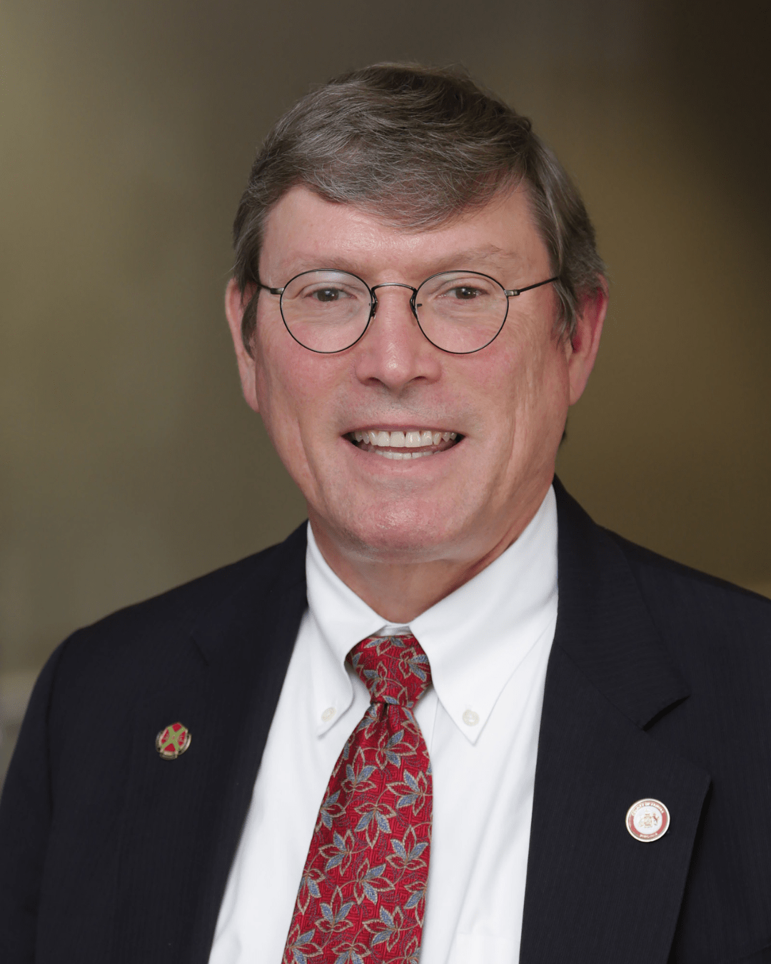 A man smiles in a headshot photo. He has short brown hair and wears glasses. He is wearing a white button-down shirt with a red tie and a black suit jacket.