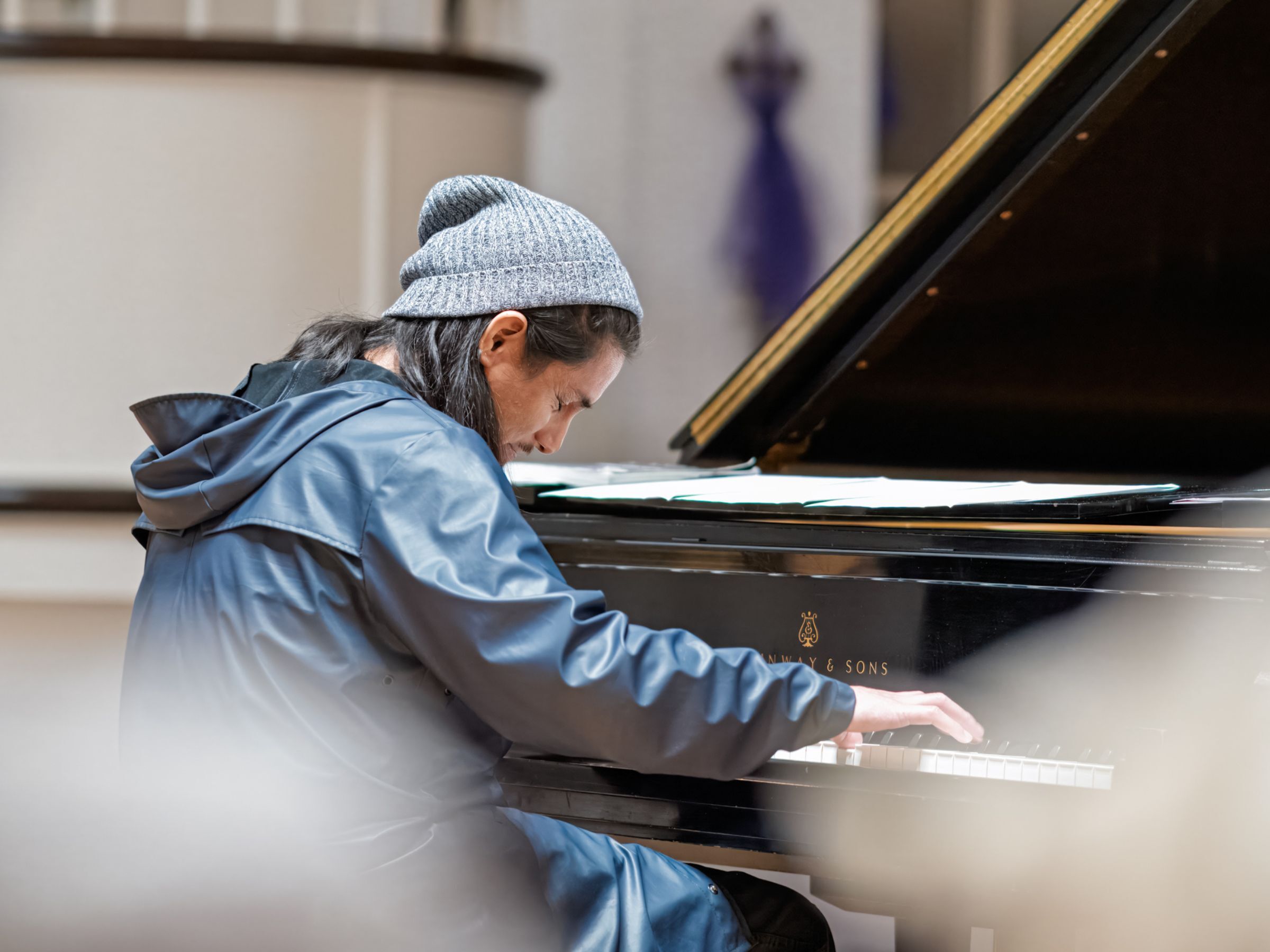 A pianist in a gray knit hat and dark gray windbreaker plays a glossy grand piano.