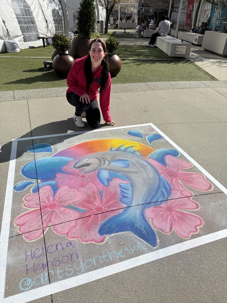 A woman with a pink jacket kneels next to her chalk art of a vibrantly colored striped bass on a paved plaza at Tysons Corner Center.