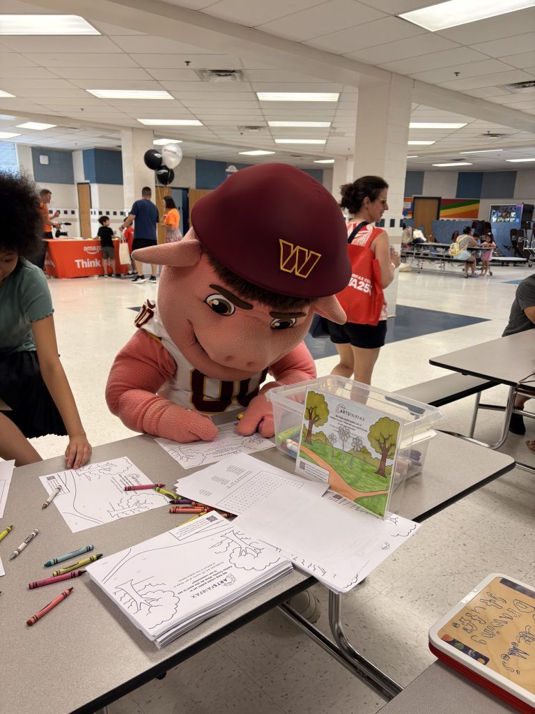 Washington Commanders mascot "Major Tuddy" is coloring an ArtsFairfax worksheet at a table.
