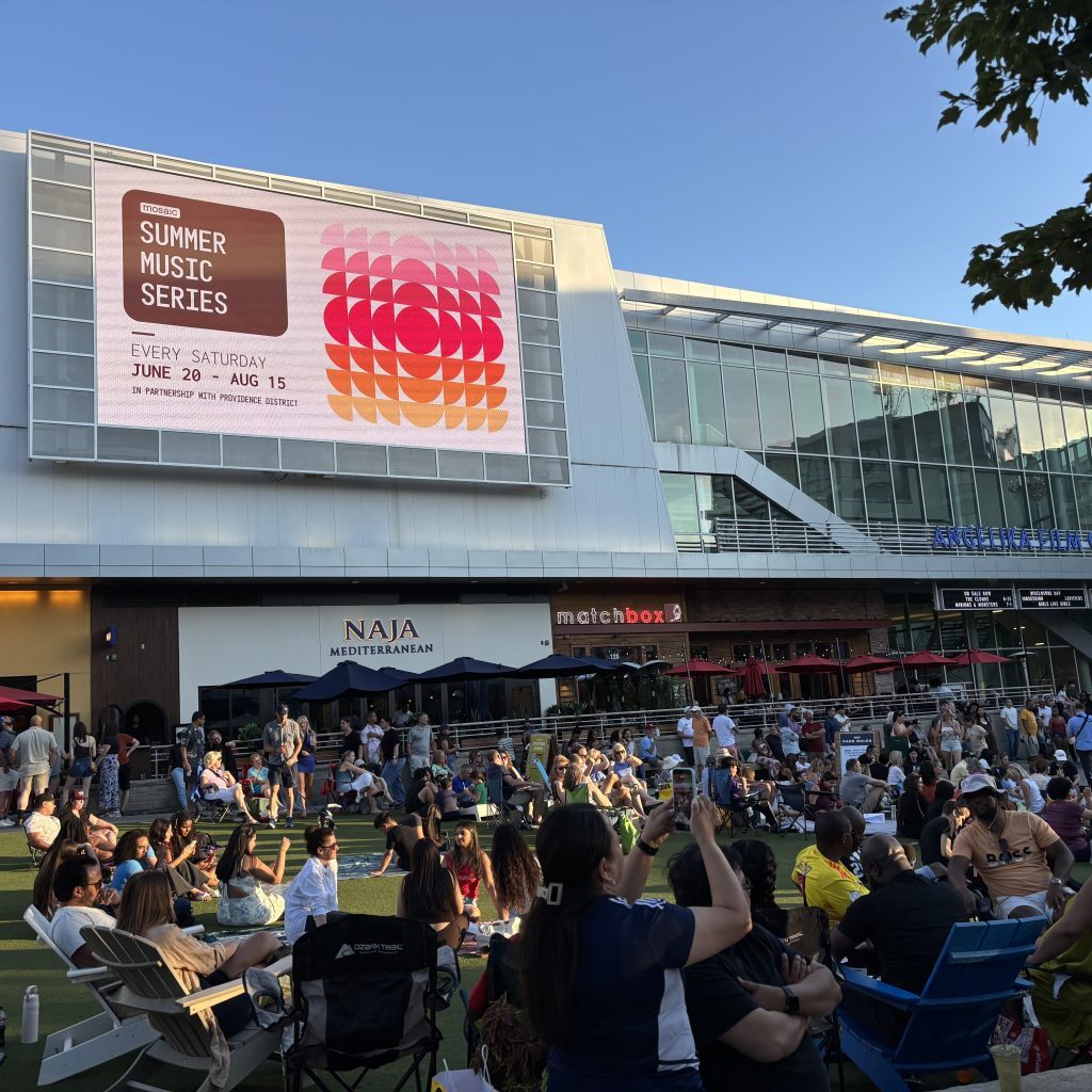 In the foreground is a lawn full of people and in the background a large screen that says "Summer Concert Series" with a pink and orange graphic.