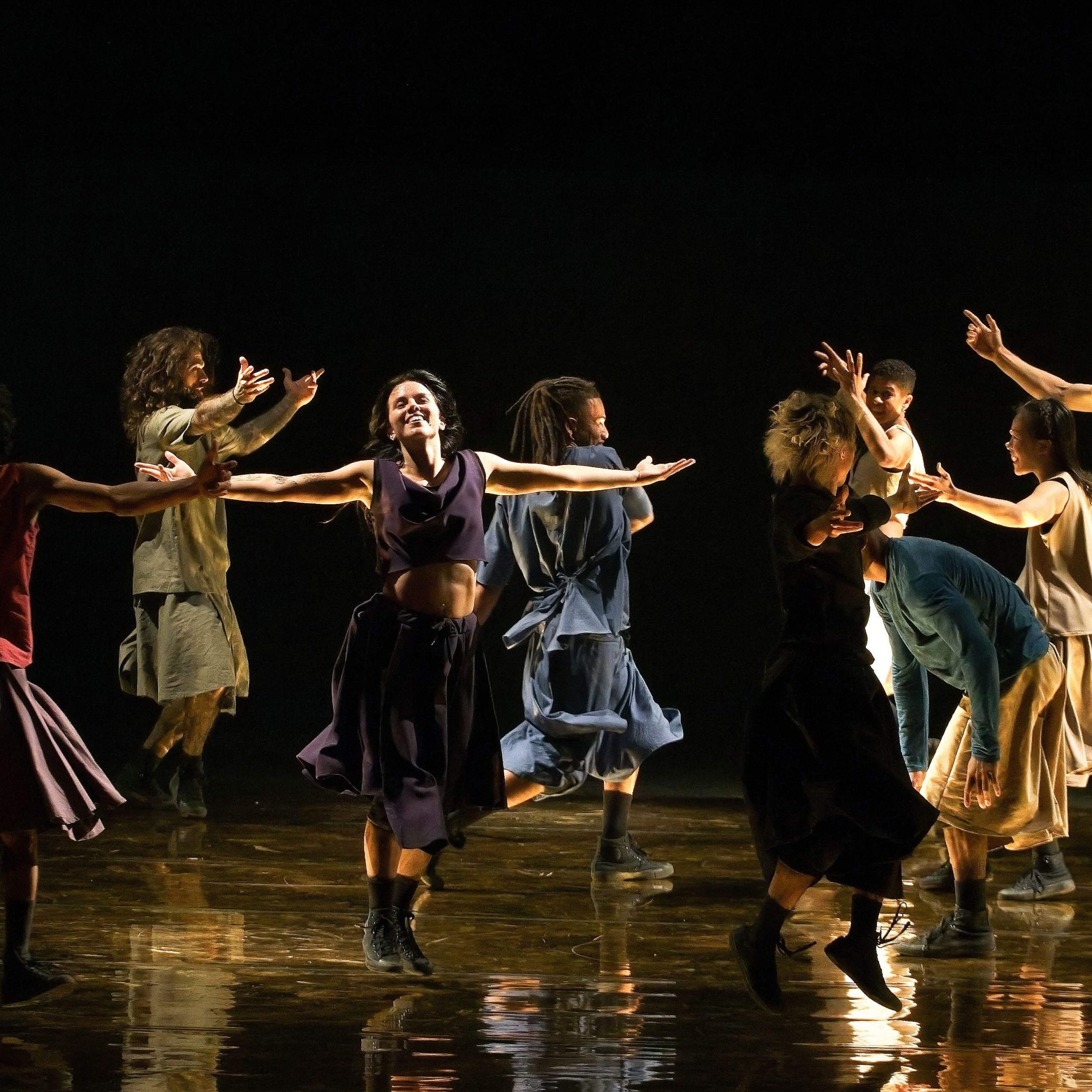 Dancers from Compagnie Hervé Koubi in loose-fitting skirts and tunics, leaping and spinning in dynamic, expressive postures across a dark stage during a live performance.