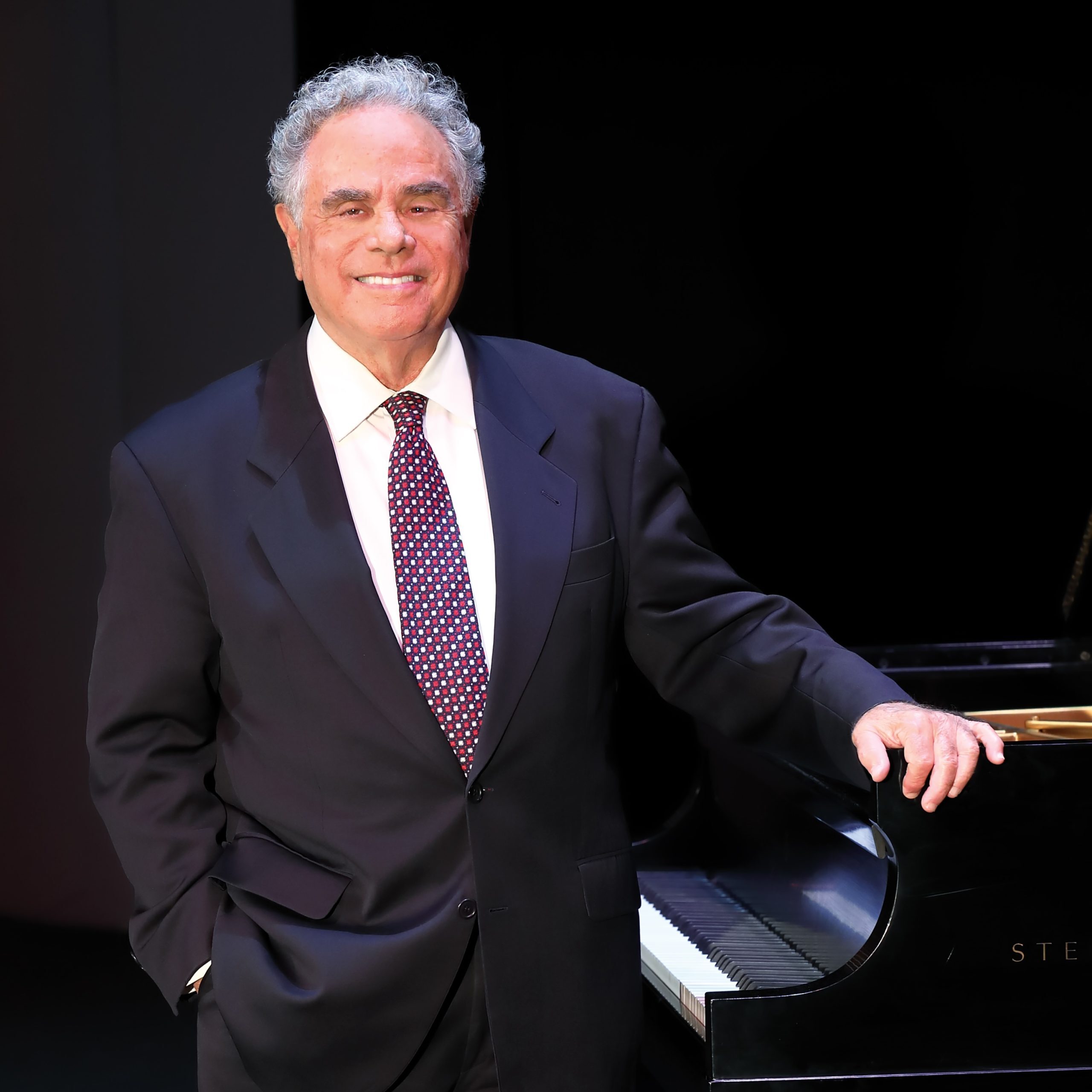 Pianist Jeffrey Siegel, in a dark suit, stands smiling with one hand resting on a black grand piano against a dark background.