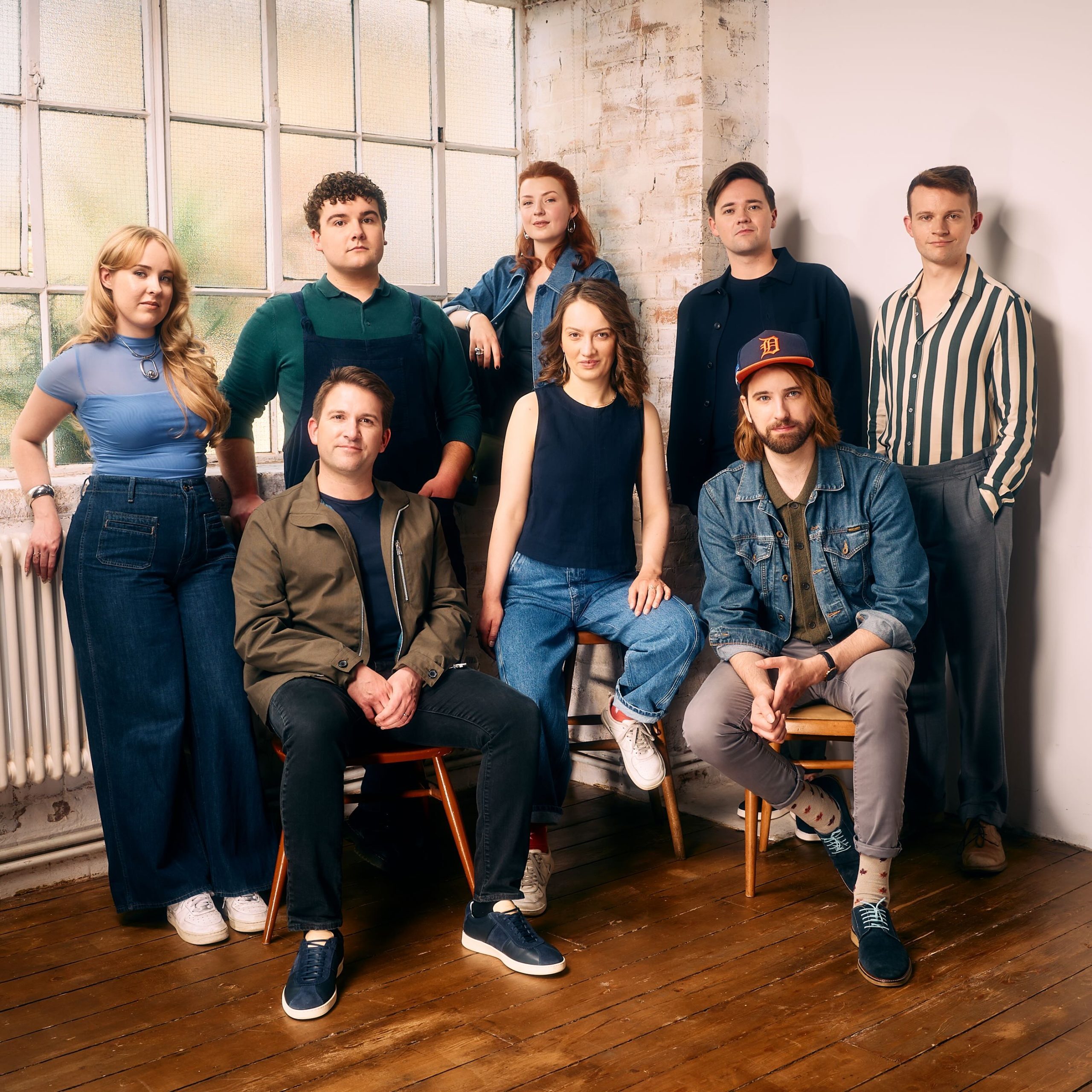 Group portrait of the eight members of VOCES8, seated and standing together in stylish denim and dark casual wear in a brightly lit studio room.