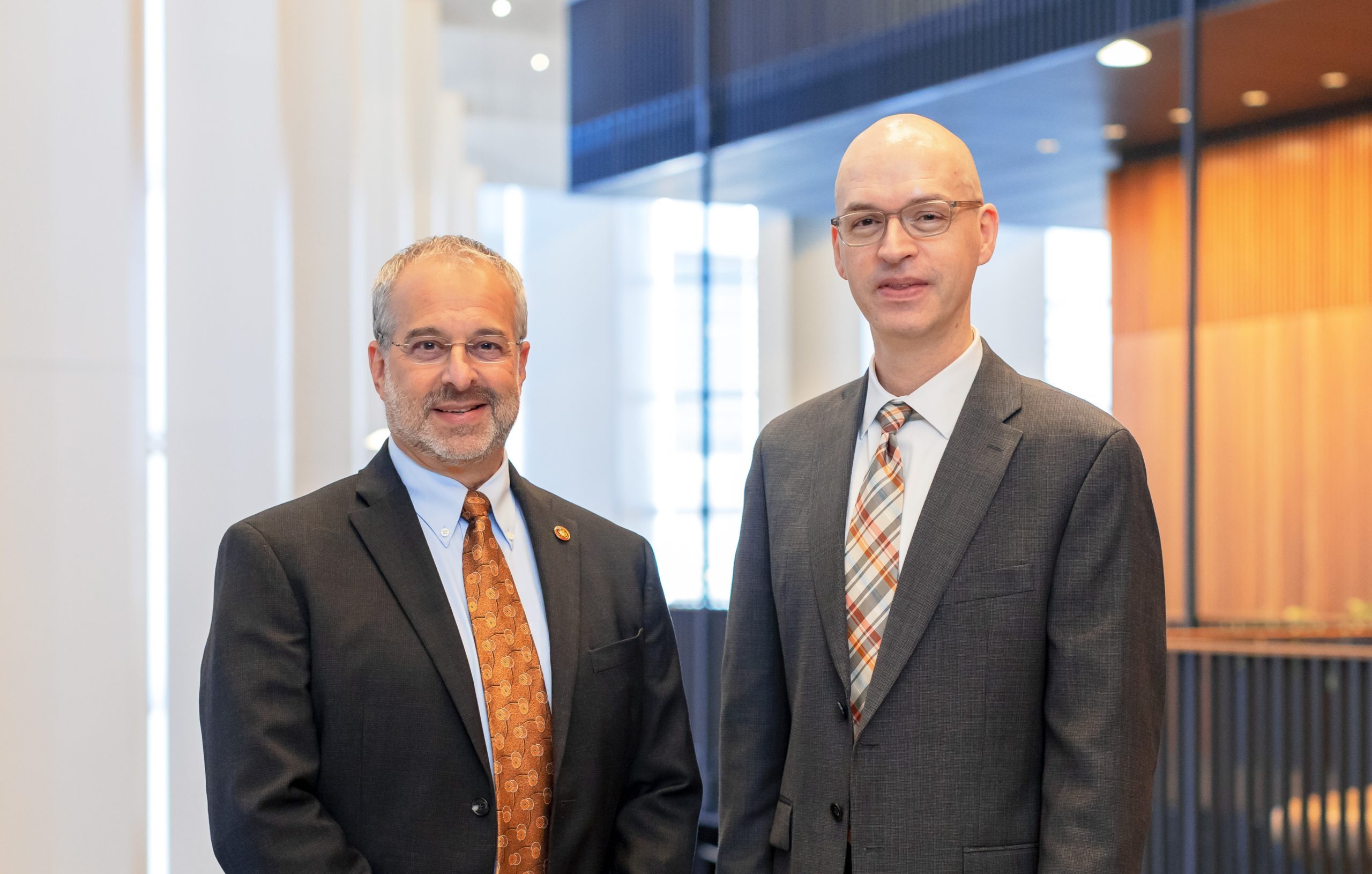 A man with salt and pepper hair, glasses in a suit and orange tie stands next to a man with translucent glaces and a multicolored tie with a brightly lit room in the background